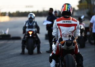 A motorcyclist wearing a vibrant racing suit with brand logos, including Repsol and Honda, sits on a sportbike on what appears to be a racetrack. In the background, another motorcyclist and a few people are present, one holding a clipboard, suggesting an organized event or race. The scene is set against a blurred backdrop, possibly indicating a focus on the individuals and the motorcycles.
