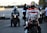 A motorcyclist wearing a vibrant racing suit with brand logos, including Repsol and Honda, sits on a sportbike on what appears to be a racetrack. In the background, another motorcyclist and a few people are present, one holding a clipboard, suggesting an organized event or race. The scene is set against a blurred backdrop, possibly indicating a focus on the individuals and the motorcycles.