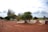 A dirt pathway leads past a cemetery dotted with gravestones and trees, with a large statue of a dog sitting prominently on the right side. In the background, a cloudy sky stretches across the horizon, and a few people stand nearby observing the scene.