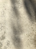 A textured concrete surface with various shades of gray and brown. The surface has darker patches, likely from stains or wear. It also shows shadows cast on it, adding depth to the flat surface.
