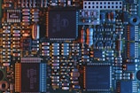 A close-up view of a computer circuit board featuring a variety of electronic components such as microchips, resistors, and capacitors. The board is highlighted with a mix of blue and orange lighting, emphasizing the intricate details and connections.