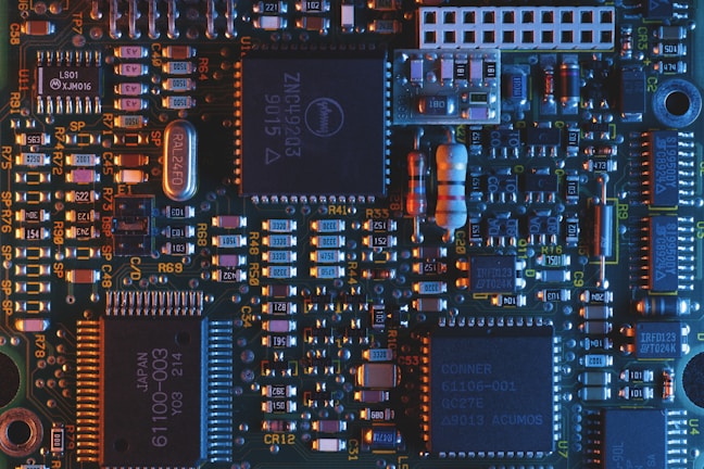 A close-up view of a computer circuit board featuring a variety of electronic components such as microchips, resistors, and capacitors. The board is highlighted with a mix of blue and orange lighting, emphasizing the intricate details and connections.