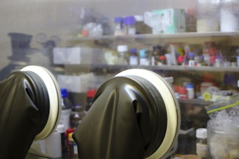 A laboratory setting featuring a pair of black gloves attached to a glass enclosure. In the background, there are shelves filled with various containers, bottles, and lab supplies, suggesting a research or experimental environment.