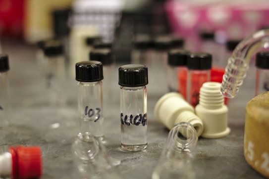 Several small transparent vials with black caps and handwritten labels are placed on a table. Around them are various lab equipment items, including plastic connectors and tubing. The background is slightly blurred, with hints of other lab materials and containers.