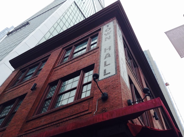 A tall, historic red brick building with multiple windows and a sign that reads 'Town Hall' in white bricks. Surrounding the structure are modern skyscrapers with reflective glass facades. The scene captures an interesting contrast between old and new architecture.