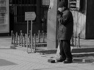 An elderly man stands on a paved sidewalk, wearing a coat and a beanie, appearing contemplative or cold. He is positioned next to a metal railing and a sign. Beside him on the ground, there is a small open container, possibly for collecting donations. The scene has a grayscale tone, emphasizing the urban setting.