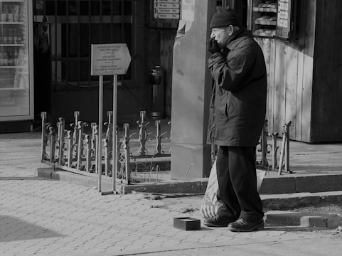 An elderly man stands on a paved sidewalk, wearing a coat and a beanie, appearing contemplative or cold. He is positioned next to a metal railing and a sign. Beside him on the ground, there is a small open container, possibly for collecting donations. The scene has a grayscale tone, emphasizing the urban setting.