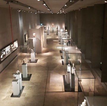 A long corridor in a museum featuring sculptures and artifacts displayed in glass cases. The room is dimly lit with spotlights highlighting each exhibit. The walls and floor have a neutral, stone-like texture, creating a calm and contemplative atmosphere.