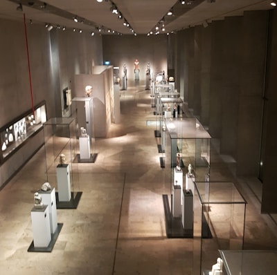 A long corridor in a museum featuring sculptures and artifacts displayed in glass cases. The room is dimly lit with spotlights highlighting each exhibit. The walls and floor have a neutral, stone-like texture, creating a calm and contemplative atmosphere.