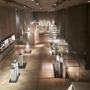 A long corridor in a museum featuring sculptures and artifacts displayed in glass cases. The room is dimly lit with spotlights highlighting each exhibit. The walls and floor have a neutral, stone-like texture, creating a calm and contemplative atmosphere.