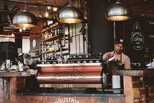 A barista prepares coffee behind a large espresso machine in a rustic, industrial-themed café. The interior features exposed brick walls, hanging pendant lights, and shelves stocked with bottles and coffee supplies. A sign on the wall reads 'Industrial Coffee.' The barista is focused on steaming milk, with various coffee-making tools placed around the counter.