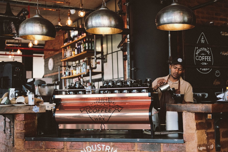 A barista prepares coffee behind a large espresso machine in a rustic, industrial-themed café. The interior features exposed brick walls, hanging pendant lights, and shelves stocked with bottles and coffee supplies. A sign on the wall reads 'Industrial Coffee.' The barista is focused on steaming milk, with various coffee-making tools placed around the counter.
