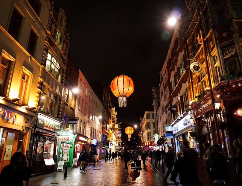 A bustling street scene at night with a vibrant atmosphere. The street is flanked by buildings featuring brightly lit signs and shopfronts. Hanging above are large, illuminated, orange lanterns that add a festive ambiance. Pedestrians and shoppers are walking along the wet pavement, which reflects the colorful lights from the shops.