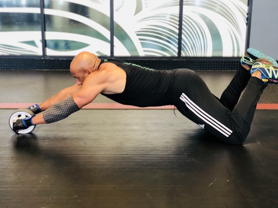 A person is performing an exercise using an ab roller in a gym setting. The individual is on their knees on a black mat, stretching forward while holding the handles of the ab roller. They are wearing a black tank top, black athletic pants with white stripes, and blue sneakers. The background features abstract patterns and a large window.