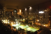 A nighttime cityscape featuring illuminated skyscrapers and high-rise buildings, with bright lights reflecting on their windows. In the foreground, several well-lit sports fields are visible, surrounded by trees and paths. The overall scene is vibrant with contrasting lights and shadows, creating a bustling urban atmosphere.