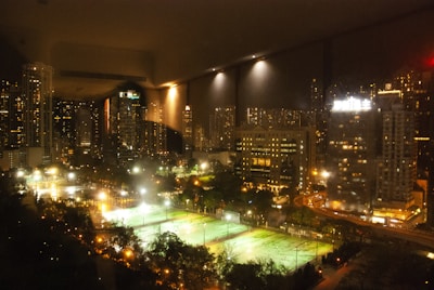 A nighttime cityscape featuring illuminated skyscrapers and high-rise buildings, with bright lights reflecting on their windows. In the foreground, several well-lit sports fields are visible, surrounded by trees and paths. The overall scene is vibrant with contrasting lights and shadows, creating a bustling urban atmosphere.