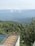 A wooden deck with a glass railing overlooks a lush, green forest. The background features misty mountains partly obscured by clouds, creating a serene and expansive landscape.
