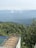 A wooden deck with a glass railing overlooks a lush, green forest. The background features misty mountains partly obscured by clouds, creating a serene and expansive landscape.