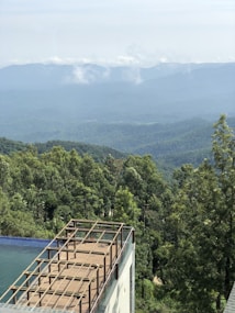 A wooden deck with a glass railing overlooks a lush, green forest. The background features misty mountains partly obscured by clouds, creating a serene and expansive landscape.