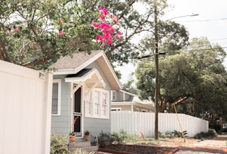 A small house with light gray exterior walls, white trim, and a small porch is surrounded by well-maintained green foliage and a white fence. There are vibrant pink flowers hanging from a tree branch in the foreground. The sky is partly cloudy, and there are utility poles and wires visible along the street.