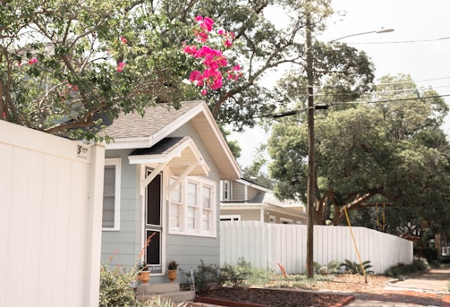 A small house with light gray exterior walls, white trim, and a small porch is surrounded by well-maintained green foliage and a white fence. There are vibrant pink flowers hanging from a tree branch in the foreground. The sky is partly cloudy, and there are utility poles and wires visible along the street.