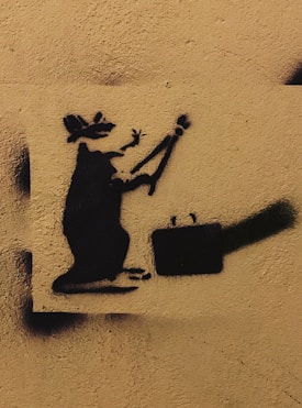 A black stencil graffiti of a rat holding a paintbrush, accompanied by a suitcase, is painted on a textured, beige wall.