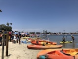 The beach scene is vibrant with numerous colorful kayaks lined up on the sandy shore. People are gathered along the beach, some in swimwear, enjoying various activities both on land and in the water. There are umbrellas and tents providing shade, and palm trees are visible in the background against a clear blue sky.