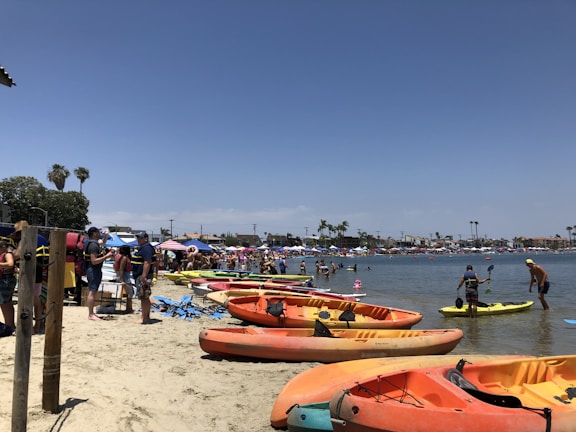 The beach scene is vibrant with numerous colorful kayaks lined up on the sandy shore. People are gathered along the beach, some in swimwear, enjoying various activities both on land and in the water. There are umbrellas and tents providing shade, and palm trees are visible in the background against a clear blue sky.