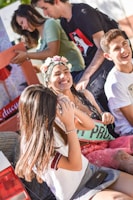 A group of young people are enjoying a fun, outdoor celebration. One person, wearing a floral headband and a sash, is smiling and looks to be the center of the event, possibly celebrating a special occasion. Others around are laughing and interacting cheerfully.