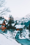 A serene alpine village nestled in the mountains during winter. Snow-covered wooden chalets are surrounded by pine trees, and vehicles are parked along a snowy road. The mountains in the background are capped with snow, and the sky is clear.