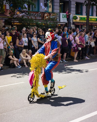 A clown with colorful makeup rides a scooter decorated with shiny yellow streamers down a street during what appears to be a parade. The crowd, consisting of adults and children, watches the performance with interest. The background shows various storefronts and a festive atmosphere.