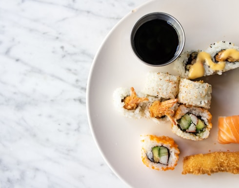 A variety of sushi rolls are arranged on a white plate, accompanied by a small metal cup of soy sauce. The sushi includes pieces with tempura, cream cheese, and cucumber, as well as a piece of salmon nigiri and a fried tempura strip.