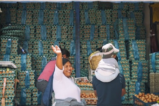 A bustling market scene with a woman smiling and raising her hand in front of a large stack of egg cartons. Three people are visible, with one carrying a child on their back. The eggs are organized in blue and green trays, creating a colorful backdrop.