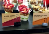 Several cups of Greek yoghurt with granola and various toppings including strawberries, raspberries, banana, and pecans are displayed. The cups are labeled as 'Mixed Berry' and 'Banana + Pecan + Chocolate' and are marked as gluten-free and sugar-free. The cups are arranged on a black surface with tags held by black binder clips.