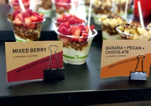 Several cups of Greek yoghurt with granola and various toppings including strawberries, raspberries, banana, and pecans are displayed. The cups are labeled as 'Mixed Berry' and 'Banana + Pecan + Chocolate' and are marked as gluten-free and sugar-free. The cups are arranged on a black surface with tags held by black binder clips.