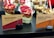 Several cups of Greek yoghurt with granola and various toppings including strawberries, raspberries, banana, and pecans are displayed. The cups are labeled as 'Mixed Berry' and 'Banana + Pecan + Chocolate' and are marked as gluten-free and sugar-free. The cups are arranged on a black surface with tags held by black binder clips.