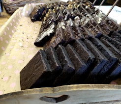 Several rows of dark chocolate brownies arranged on a pink polka-dotted paper-lined wooden tray. The brownies are neatly stacked, with some topped with different kinds of nuts and drizzled chocolate.