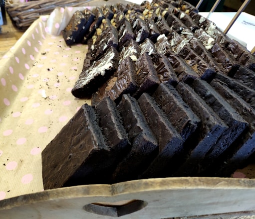 Several rows of dark chocolate brownies arranged on a pink polka-dotted paper-lined wooden tray. The brownies are neatly stacked, with some topped with different kinds of nuts and drizzled chocolate.