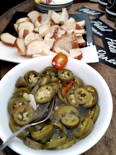 A bowl filled with slices of pickled jalapeños is centered in the foreground, with some red peppers mixed in. To the side in the background, there is a platter of sliced bread. The setting includes other items like jars and plates arranged on a wooden surface.
