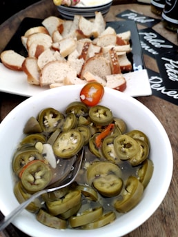 A bowl filled with slices of pickled jalapeños is centered in the foreground, with some red peppers mixed in. To the side in the background, there is a platter of sliced bread. The setting includes other items like jars and plates arranged on a wooden surface.