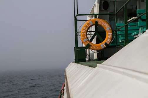 A foggy scene on a maritime vessel, showing an orange life ring mounted on the green railing. The ship's structure features metal and wooden parts and the ocean is visible in the background.