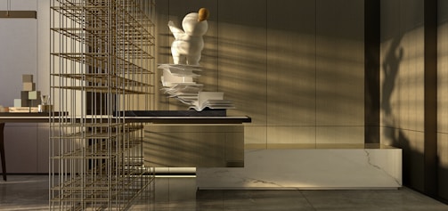 A modern interior space with a focus on minimalism. There is a large, abstract sculpture resembling a person or character with spherical forms, positioned on a surface with open books scattered alongside. The sunlight casts shadows across the room, highlighting the neutral tones and sleek, contemporary design. A decorative grid structure adds an element of intrigue on the left side. The materials used include marble and wood, contributing to the sophisticated atmosphere.
