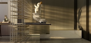 A modern interior space with a focus on minimalism. There is a large, abstract sculpture resembling a person or character with spherical forms, positioned on a surface with open books scattered alongside. The sunlight casts shadows across the room, highlighting the neutral tones and sleek, contemporary design. A decorative grid structure adds an element of intrigue on the left side. The materials used include marble and wood, contributing to the sophisticated atmosphere.