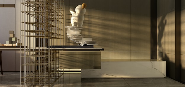 A modern interior space with a focus on minimalism. There is a large, abstract sculpture resembling a person or character with spherical forms, positioned on a surface with open books scattered alongside. The sunlight casts shadows across the room, highlighting the neutral tones and sleek, contemporary design. A decorative grid structure adds an element of intrigue on the left side. The materials used include marble and wood, contributing to the sophisticated atmosphere.