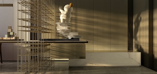 A modern interior space with a focus on minimalism. There is a large, abstract sculpture resembling a person or character with spherical forms, positioned on a surface with open books scattered alongside. The sunlight casts shadows across the room, highlighting the neutral tones and sleek, contemporary design. A decorative grid structure adds an element of intrigue on the left side. The materials used include marble and wood, contributing to the sophisticated atmosphere.