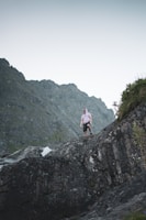 A person is hiking on a rugged rocky terrain with a mountainous backdrop. They are wearing casual clothing and appear to be engaged in an outdoor activity.