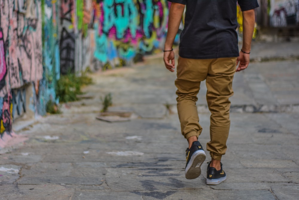 hombre vestido con camisa negra y pantalones marrones caminando junto a la pared con graffiti