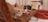 An orange tabby cat with a red and green collar is perched attentively on a wooden bench, while a second cat with a mix of black, white, and brown fur rests on a cushion behind it. The scene is indoors, with soft focus on the background, suggesting a cozy and relaxed setting.
