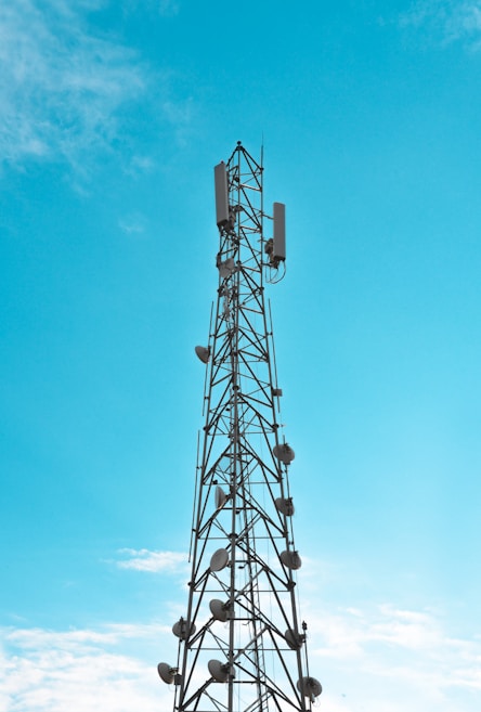 A tall telecommunications tower extends into the bright sky, adorned with numerous satellite dishes and antennae along its structure. The sky is a clear blue with a few scattered clouds.