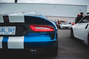 A parking lot scene with a focus on a blue sports car with white racing stripes. The rear end of the car is prominently displayed, showcasing its taillights and license plate. Other vehicles and people in the background suggest a car gathering or event.